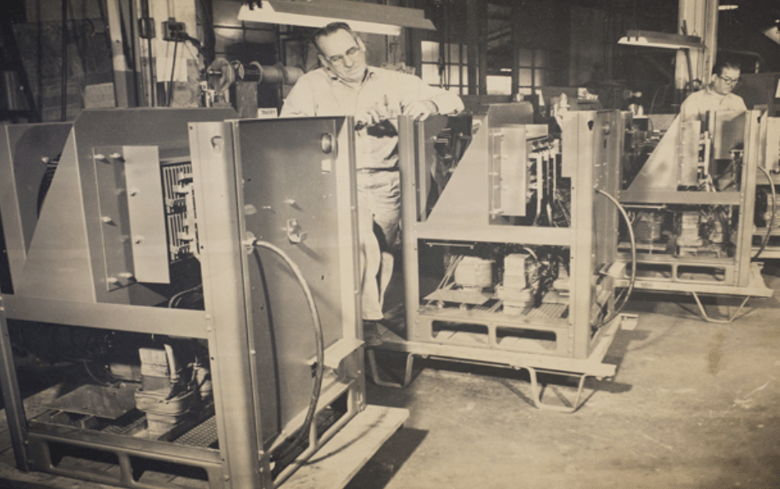 Two technicians work on machinery in a factory setting, inspecting large metal units. The image highlights manufacturing processes and technical skills.
