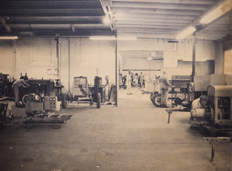 A busy workshop scene with several workers assembling machinery. Tools and equipment are visible, highlighting an industrial setting focused on production.