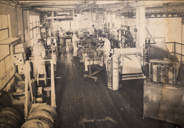 Historical workshop setting with workers in white coats, engaged in machinery assembly and engineering tasks, showcasing industry from the past.