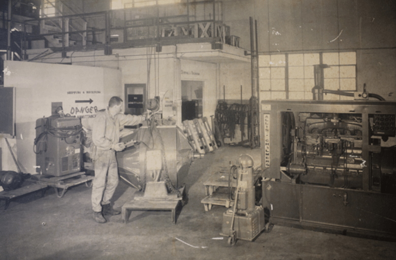 A worker inspects a large industrial machine in a factory setting. Safety warnings are visible on the walls, emphasizing caution in the workspace.